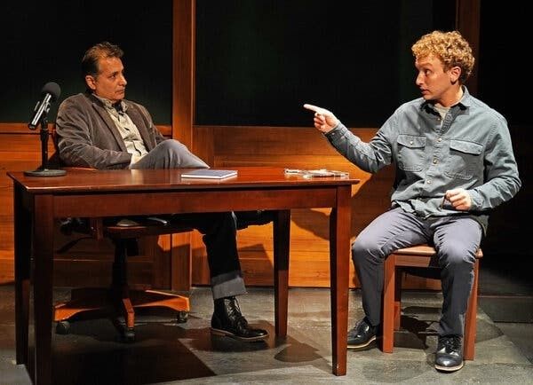 Two actors stage a dramatic dialogue across a desk