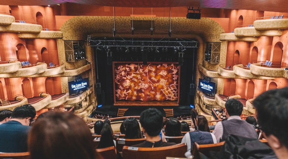 Interior view of an active theater showing a full audience watching a performance on stage, with multiple tiers of seating and professional theater lighting visible.